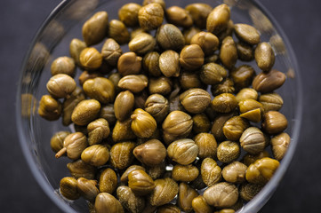 pickled capers in a glass bowl on a dark background, top view close up