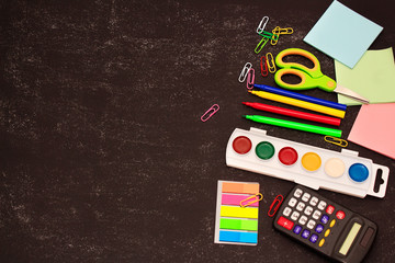 School accessories (pencils, pens, calculator, paper clips, paper, felt-tip pens, paints, scissors) on black paper background. School preparation concept