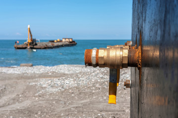 Fototapeta na wymiar An industrial rusty water faucet on the shore with blurred barge in the sea