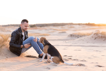 Fototapeta na wymiar A young Caucasian man dressed black leather jacket and blue jeans sits on sandy beach next to his friend the dog Beagle breed.