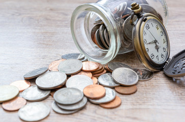 Vintage pocket watch and coins open glass jar on wooden background. Financial concept of time and saving money