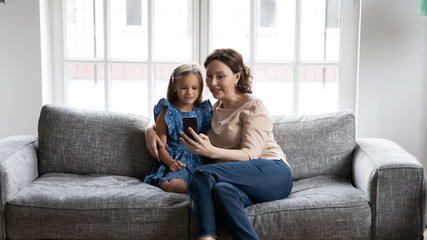Loving middle-aged grandmother and little granddaughter sit relax on sofa at home using cellphone together, caring mature granny with small girl child rest on couch watch video on smartphone gadget