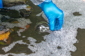 hand in a blue rubber glove pours water from a river into a transparent flask for analysis in a...