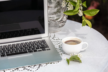 Cup of coffee, laptop, bird cherry color on the table, blurred garden background, green leaves, flowers