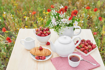 Romantic Valentines french or rural breakfast: tea, strawberries, croissants on table in poppy field. Countryside and Cozy Good morning weekend concept. Background with copy space. Date outdoors