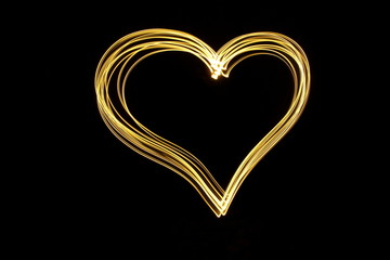 Long exposure photograph of neon gold colour in an abstract heart shape outline, parallel lines pattern against a black background. Light painting photography.