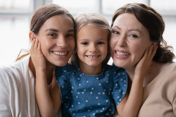 Portrait of little preschooler girl hug smiling young mother and senior grandmother, happy three generations of women embrace cuddle, enjoy family reunion, weekend at home together, bonding concept