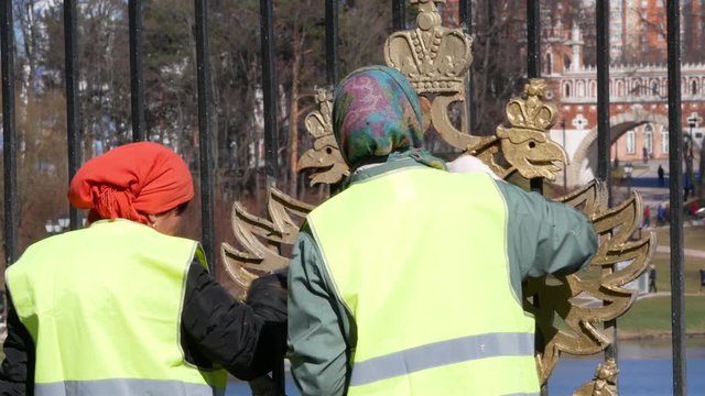 Two working women in bright vests paint the coat of arms of Russia on the fence of the park