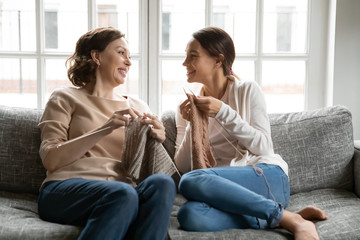 Smiling middle-aged mother and adult daughter relax on sofa at home knit with needles together,...