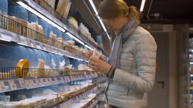 Pretty young woman choosing dairy products in the supermarket. Text on jar "robiola", on boxes "blue cheese with mold", "belgian cheese delis", "brie defamilier"