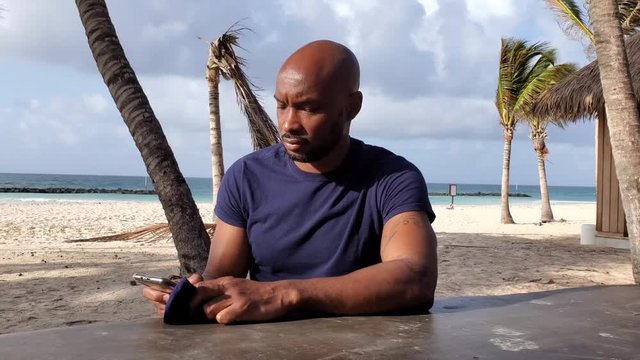 Young Black African American man reads news on mobile phone using data / WiFi then remembers to wear protective face mask as Beach reopens in the Caribbean 
