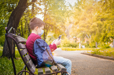 Fototapeta premium A teenager in a medical mask is sitting on a bench in an autumn park with a smartphone.