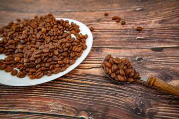 A spoon with roasted coffee lies on a wooden background, next to a white plate with coffee grains.