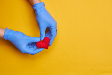 red heart symbol at persons hand with blue medical gloves on. yellow or orange background with copy space. heart disease concept.
