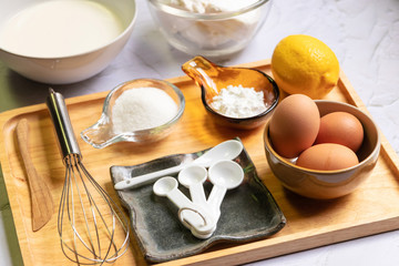 Baking ingredients for cheesecake on wooden tray. Eggs, flour, sugar, lemon, cream, creamcheese and kitchen utensils on table.