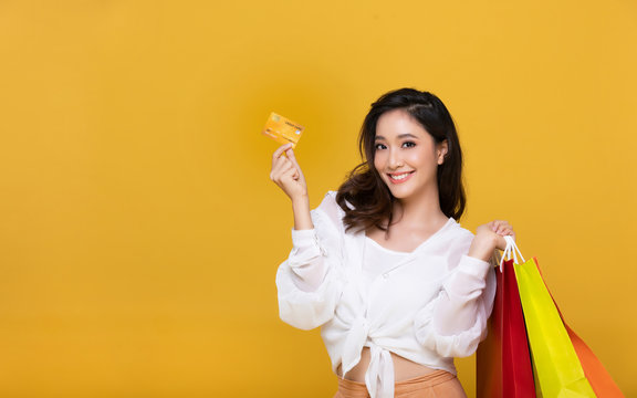 Portrait Asian beautiful happy young woman smiling cheerful and she is holding credit card and using smart phone for shopping online with shopping bags on yellow background.