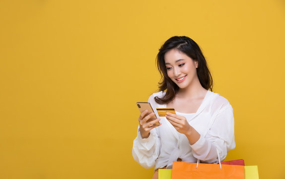 Portrait Asian beautiful happy young woman smiling cheerful and she is holding credit card and using smart phone for shopping online with shopping bags on yellow background.