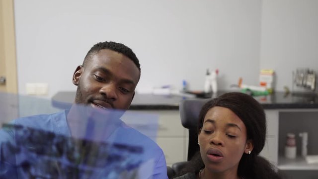 A young handsome African man dentist in a blue medical suit and blue gloves shows his female pretty patient a panoramic dental X-ray image of the jaws and talks about teeth treatment
