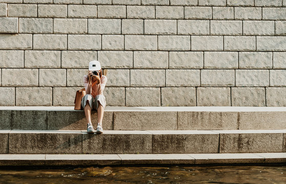 Portrait Of A Young Asian Hipster Girl With A Camera Taking Photos And Sitting On The Stairs In The City. Travel And Work Freelance Concept. Hipster Style.