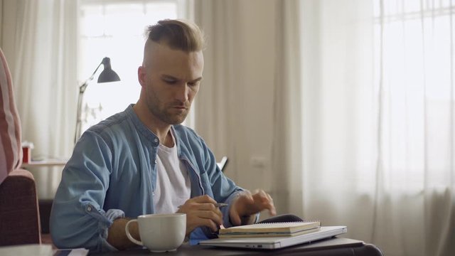 Side view of young man with coffee cup sitting down on sofa, opening laptop watching video conference and making notes in notepad, then closing laptop and day dreaming at home