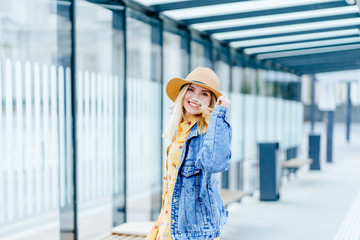 Portrait of blond positive smiling woman traveler in straw hat, denim jacket, summer dress dancing, whirling enjoying in bus stop public outdoor.