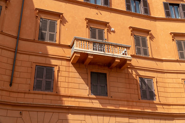 Fototapeta na wymiar View from below on building facade. A sunny stone balcony at an orange townhouse building with geometric lines in a street in Rome, Italy. Many windows closed with brown shutters