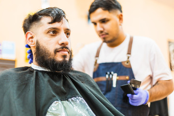 Fototapeta premium Young Latin Barber At Work In Stylish Barbershop. Cool, bearded man enjoys the moment.