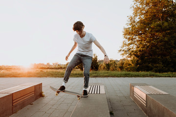 Junge moderne Frau fährt auf dem Skateboard und grinded auf der Bank im Park bei Sonnenuntergang im Sommer. Das Mädchen trägt ein weißes T-Shirt und eine blaue Jeans beim skaten und macht einen Trick. - obrazy, fototapety, plakaty