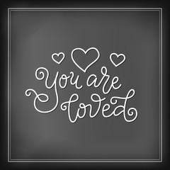 Modern mono line calligraphy lettering of You are loved in white with hearts on chalkboard