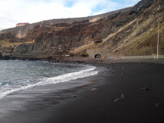 Ocean waves on a black volcanic beach. On the horizon is a brown mountain, a white house and a tunnel.