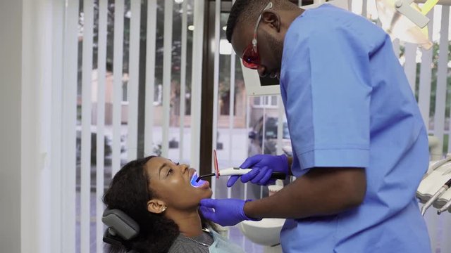 Young male African doctor dentist fixing and drying dental fillings with a special light, holding a polymerization lamp in his hands. Female patient visiting dentist for regular check-up and filling