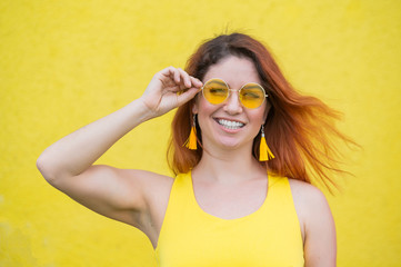 Portrait of a happy woman in sunglasses on a yellow background. Red-haired carefree girl in a dress and earrings tassels of the same color. Lady with an impeccable dazzling smile. Summer photo.