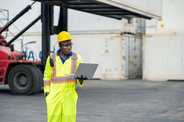 African technician dock worker in protective safety jumpsuit uniform and with hardhat and use laptop computer at cargo container shipping warehouse. transportation import,export logistic industrial