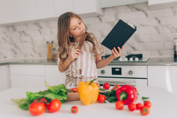 Little girl cooking and holding a recipe book in his hands in kitchen at home.