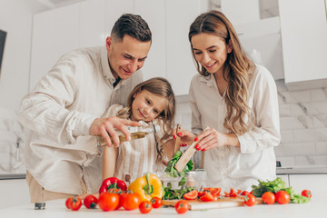 Happy family of little girl and her parents having fun and cooking salad in kitchen at home.