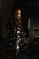 the girl sits on a chair in a plaid shirt, put her hands on the back of the chair, the ray of the sonde falls on her face