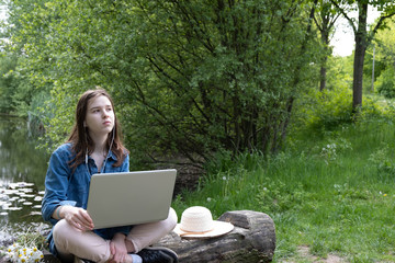 near the lake, outside the city, a young girl sits on an old log with a laptop and thinks
