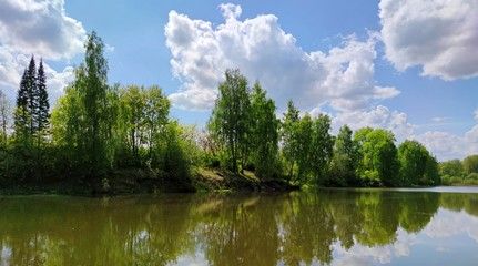 green trees by the lake are reflected in the water against a blue sky with clouds on a sunny day