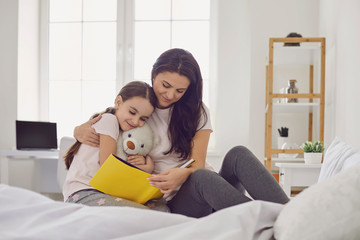 Happy Mother's Day. Mother and daughter are reading a book while sitting on the bed at home.