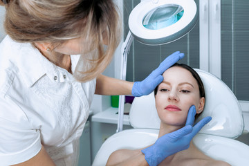 Young woman at beauty clinic cosmetology service sitting on medical chair while female doctor examining skin with mirror