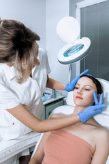 Young woman at beauty clinic cosmetology service sitting on medical chair while female doctor examining skin with mirror