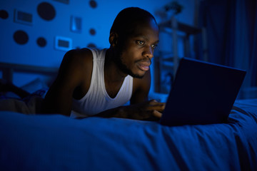 Portrait of young African-American man using laptop while lying on bed at night, copy space