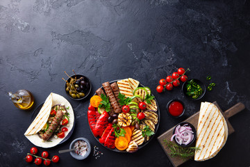 Grilled meat kebabs, vegetables on a black plate with tortillas, flat bread. Slate background. Copy...