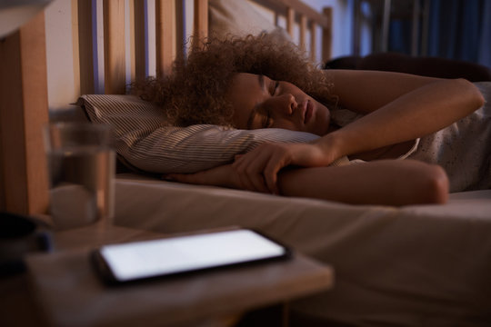 Portrait of curly-haired young woman sleeping calmly in bed at night with lit smartphone on nightstand, copy space