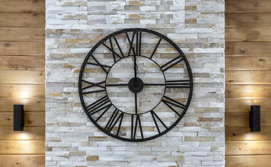 Industrial style metal clock with a Roman dial on a marble wall.