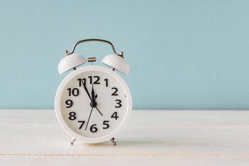 White alarm clock standing on wooden shelf on blue background. copy space fro your text or design. Fime minutes before twelve on a display