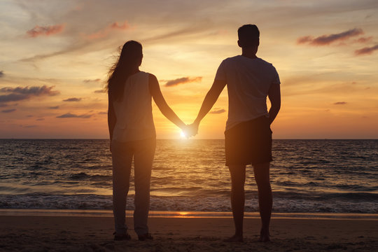 silhouette of young couple standing on beach to boundless ocean water joining hands at orange sunset backside view