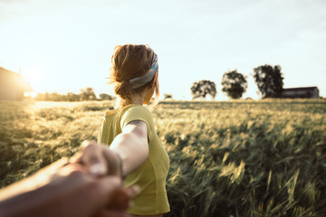 Fototapeta na wymiar Pov view of a happy young woman holding hand of her boyfriend while walking by a wheat field at sunset. Couple enjoying travel in the nature