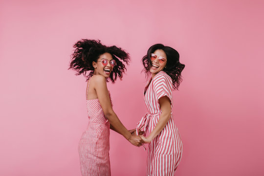 Winsome girls in glamorous glasses laughing together on pink background. Indoor photo of amazing international female friends holding hands and dancing