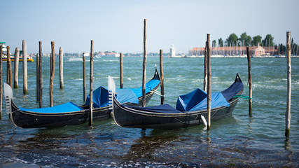 Two blue gondolas are moored on wooden pillars with lagoon on background